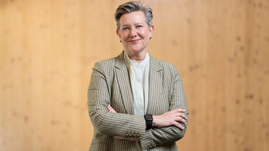 Professor Joan Leach stands with her arms crossed, smiling. She is wearing a gray blazer.